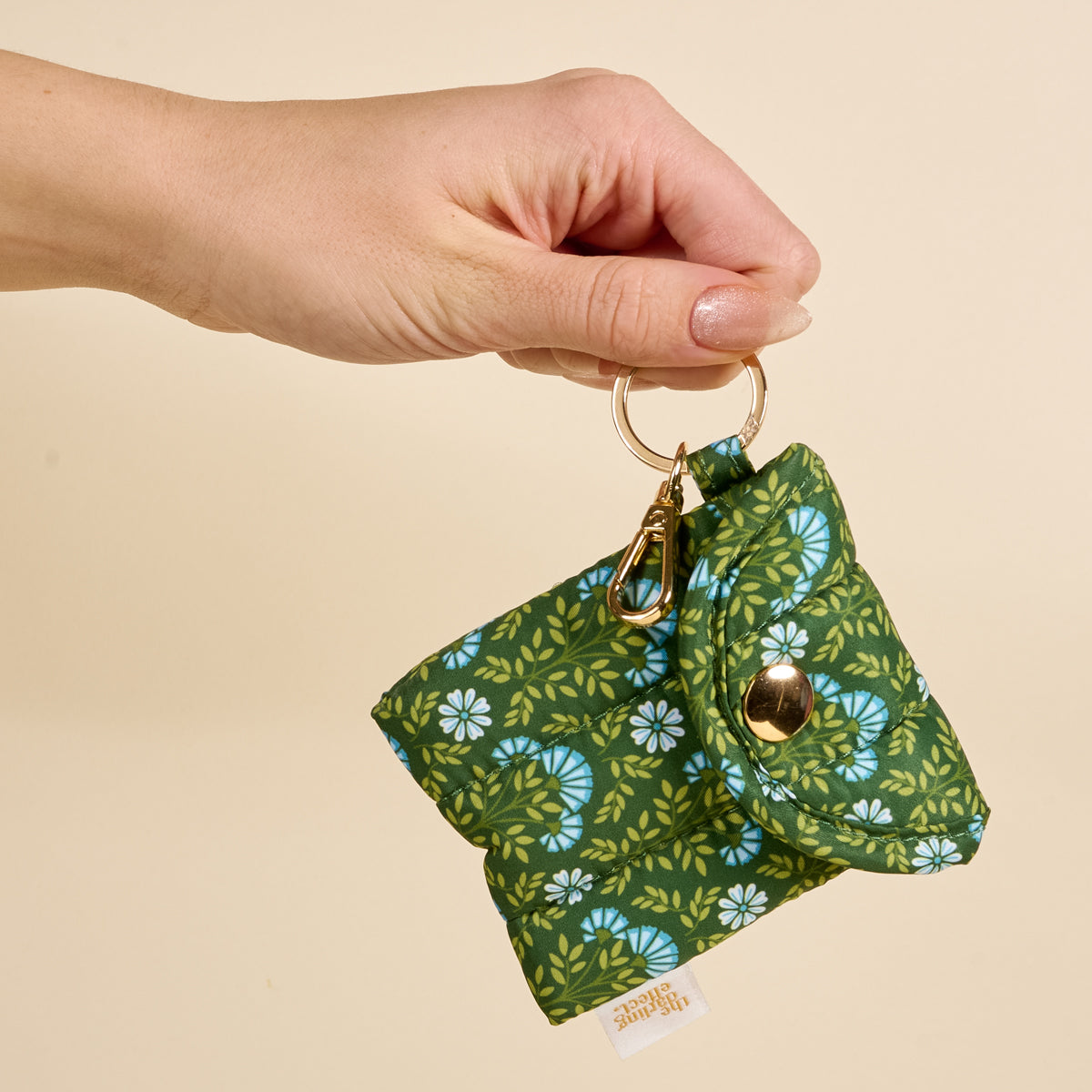 Hand holding The Darling Effect Everbloom Forest Green Puffy Earbud Case, featuring a quilted design with green floral pattern, gold snap closure, and keychain clip—ideal for wireless earbuds—against a beige background.