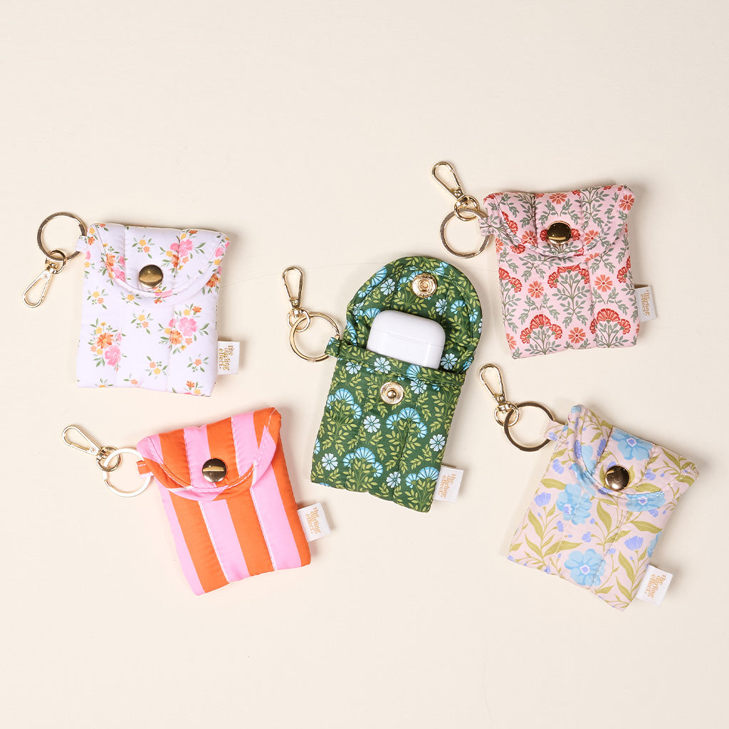 Five small, colorful Puffy Earbud Cases from The Darling Effect are arranged in a circle, each with a keychain clip. In the center, the Everbloom Forest Green Puffy Earbud Case is open to reveal a white wireless earbuds case inside.