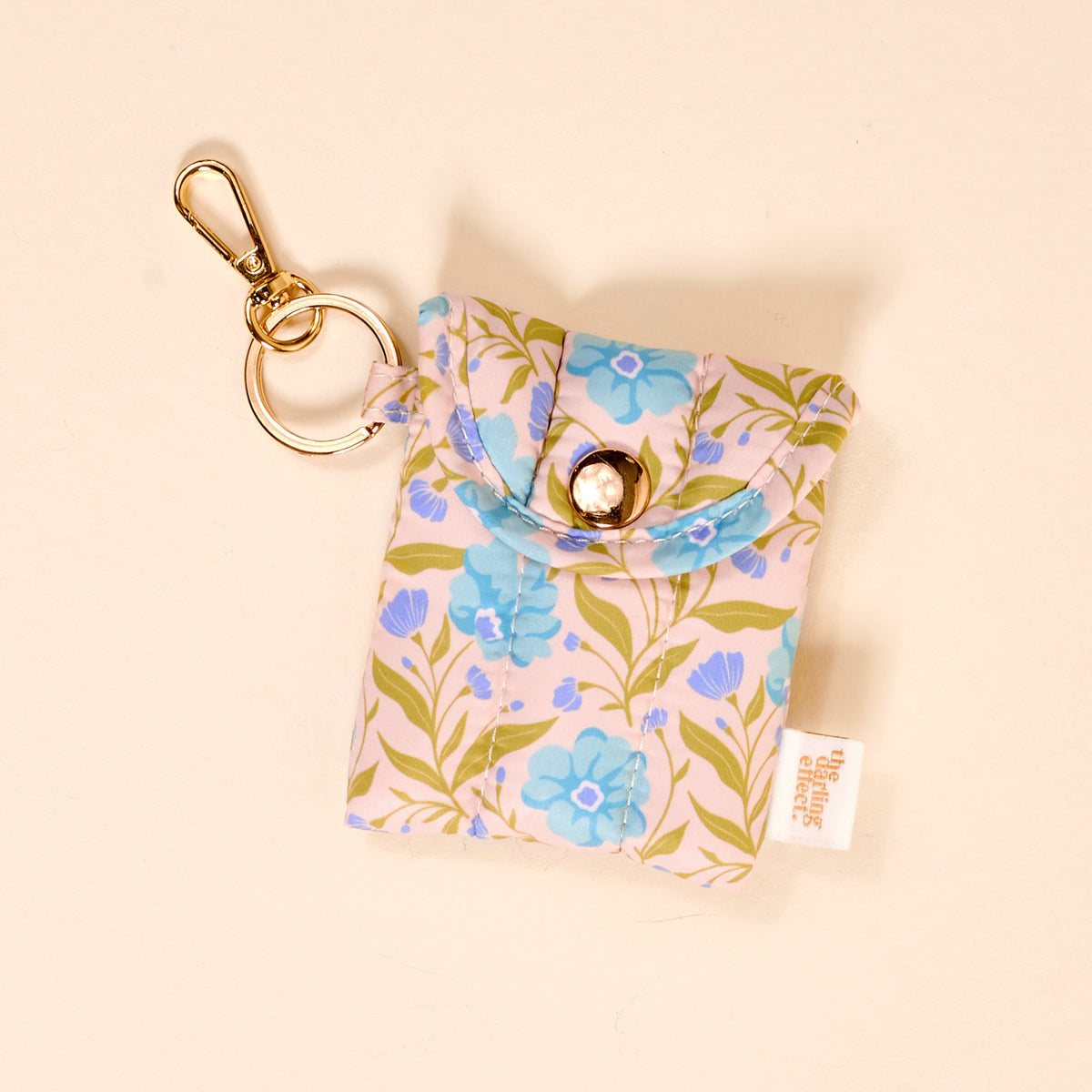 The Darling Effect’s Poppin' Petals Aqua Puffy Earbud Case is a small fabric keychain pouch with a blue and green floral pattern, gold keyring and clasp, button closure, and a white side tag.