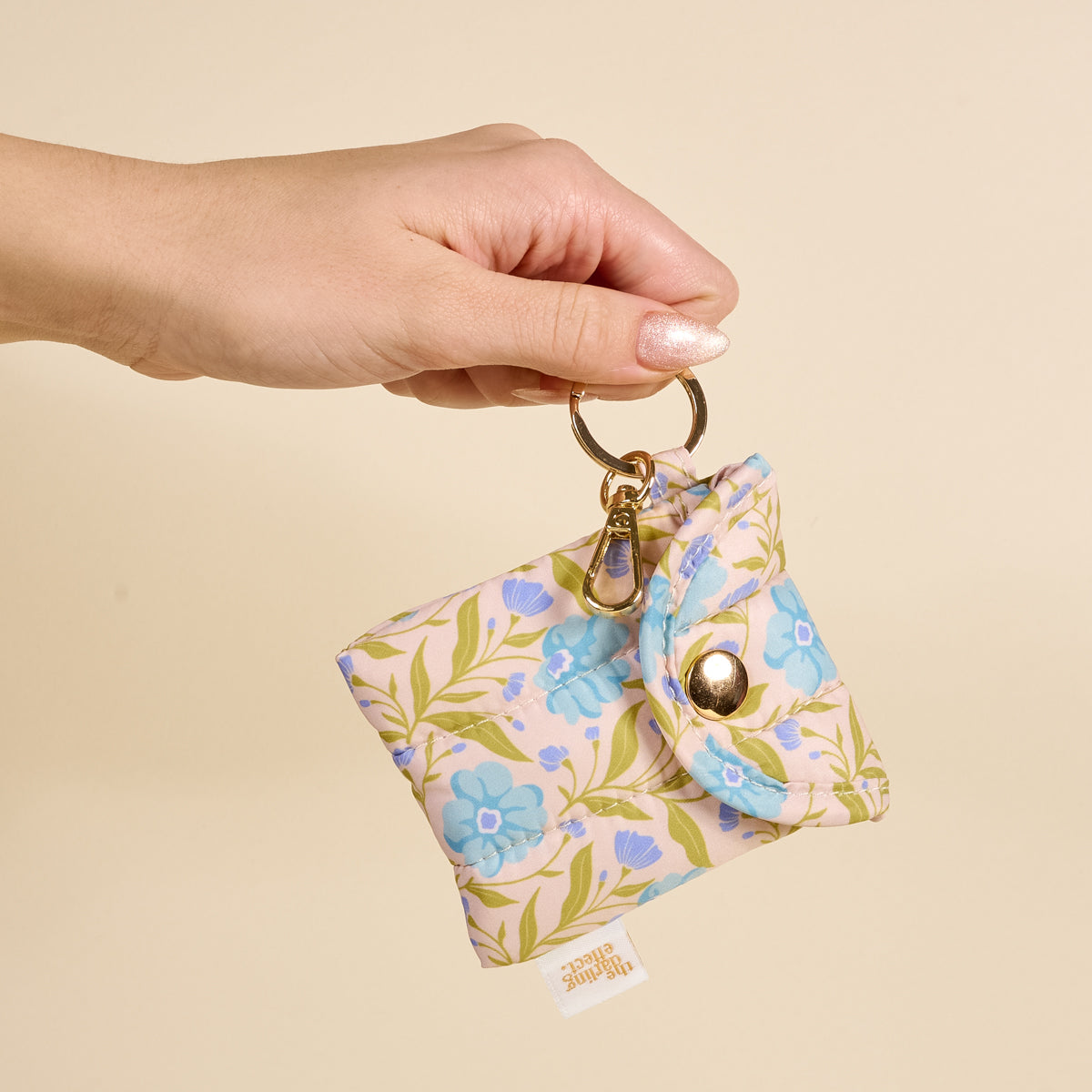 A hand holds The Darling Effect Poppin' Petals Aqua Puffy Earbud Case—a small fabric pouch for wireless earbuds, with blue and green floral print, gold keychain and clasp, and a secure button closure.