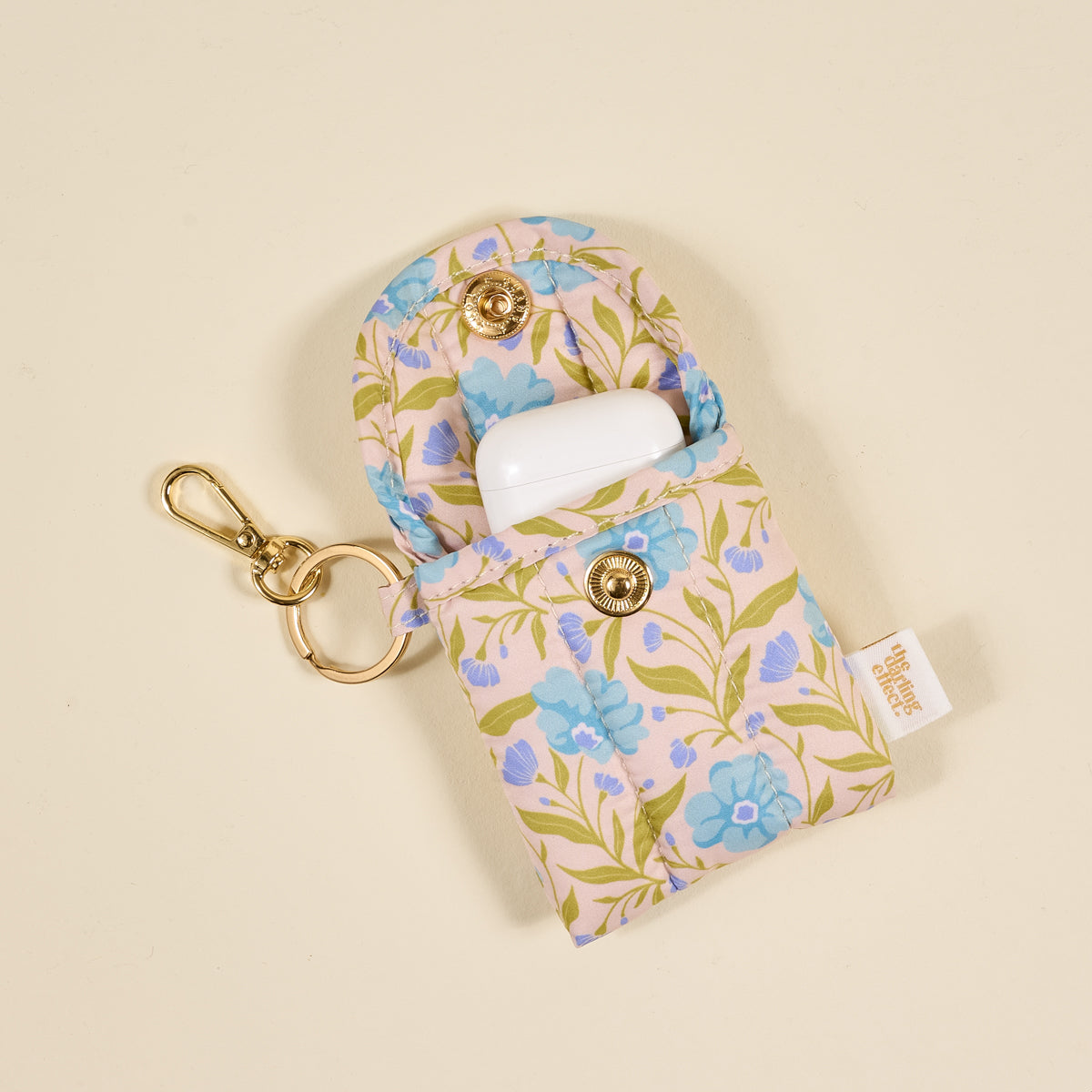 The Darling Effect's Poppin' Petals Aqua Puffy Earbud Case features a blue floral design, gold snap buttons, and a keychain. It securely holds an open white wireless earbuds case inside.