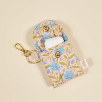 The Darling Effect's Poppin' Petals Aqua Puffy Earbud Case features a blue floral design, gold snap buttons, and a keychain. It securely holds an open white wireless earbuds case inside.