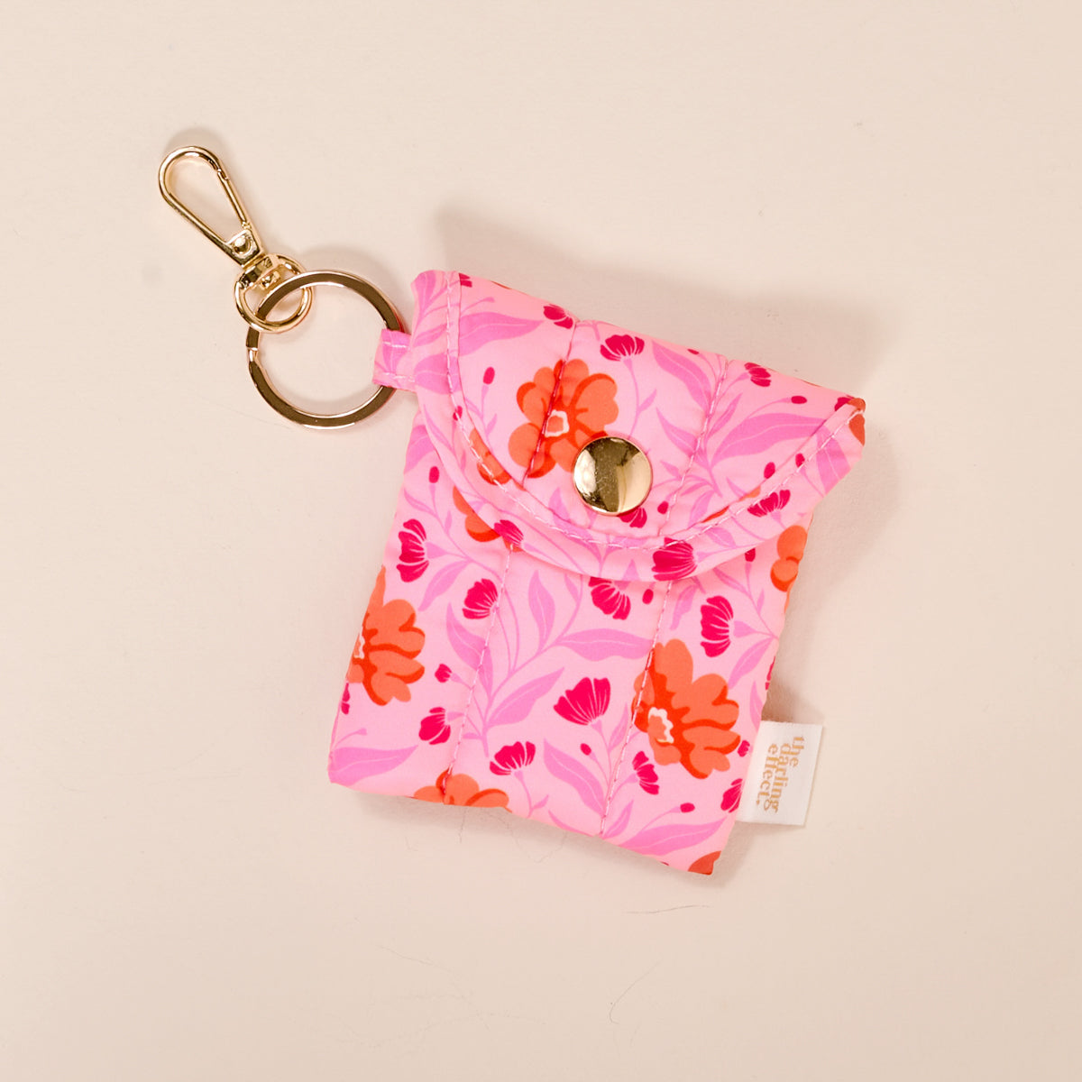 The Darling Effect's Poppin' Petals Hot Pink Puffy Earbud Case features a small, pink floral design, snap button closure, gold keychain attachment, and white fabric tag. Shown on a light background.