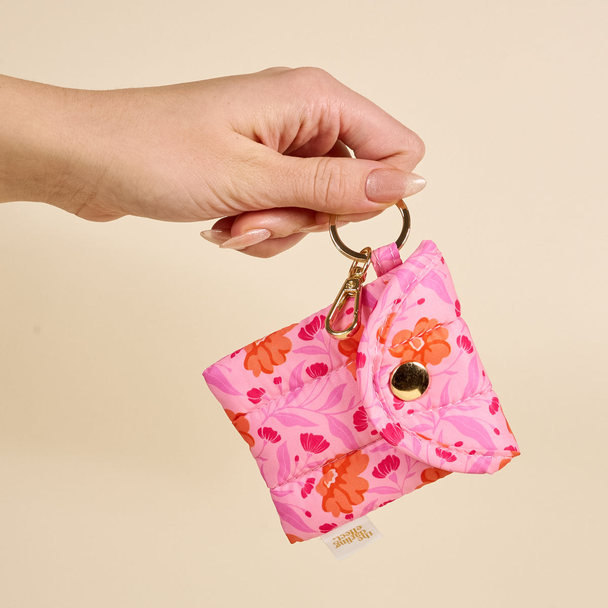 A hand holds The Darling Effect Poppin' Petals Hot Pink Puffy Earbud Case, a quilted floral-patterned pouch keychain with a gold snap button, against a beige background.