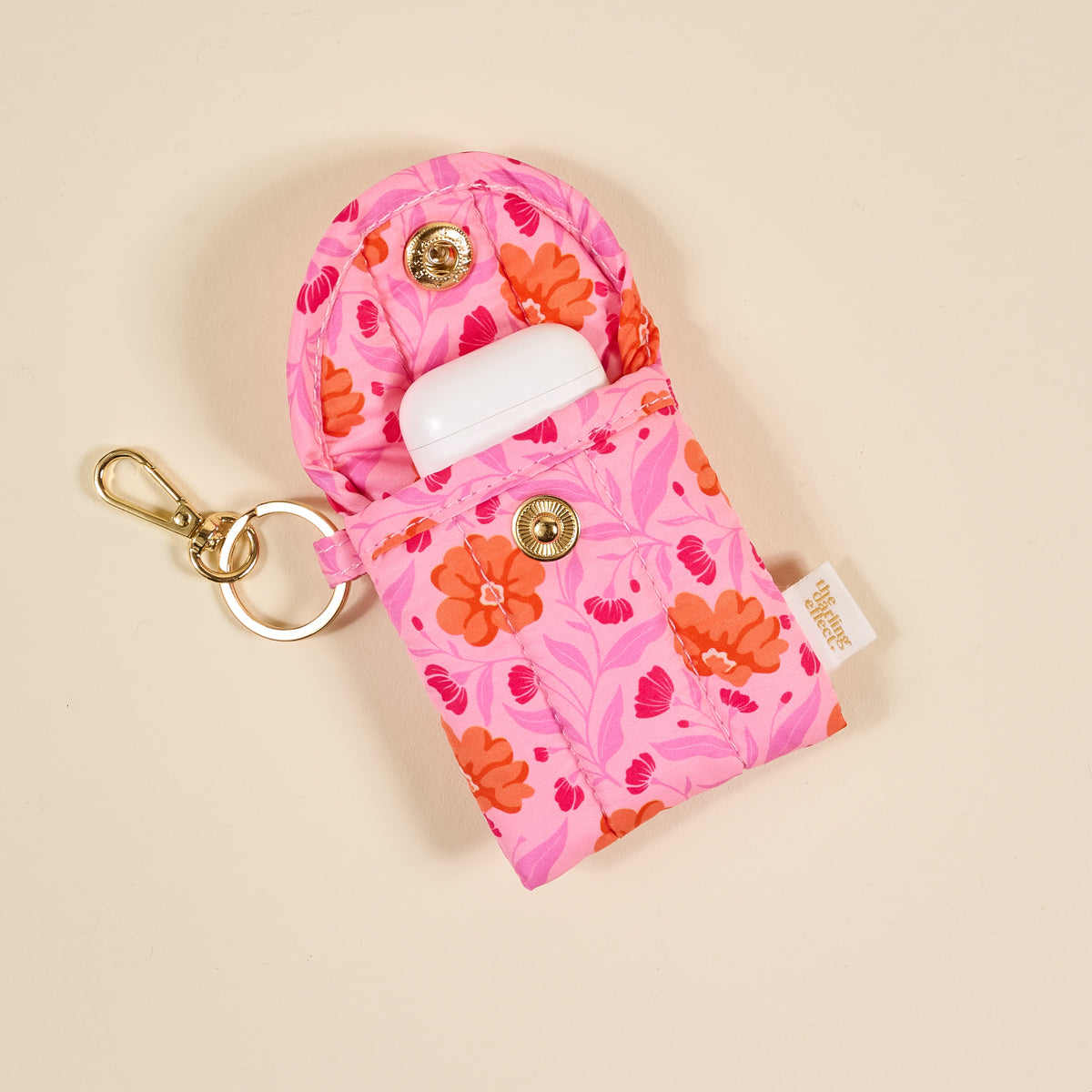 The Darling Effect’s Poppin' Petals Hot Pink Puffy Earbud Case features a pink floral design, snap button closure, gold keyring and clasp, white wireless earbuds inside, and a side fabric tag for a stylish keychain accessory.