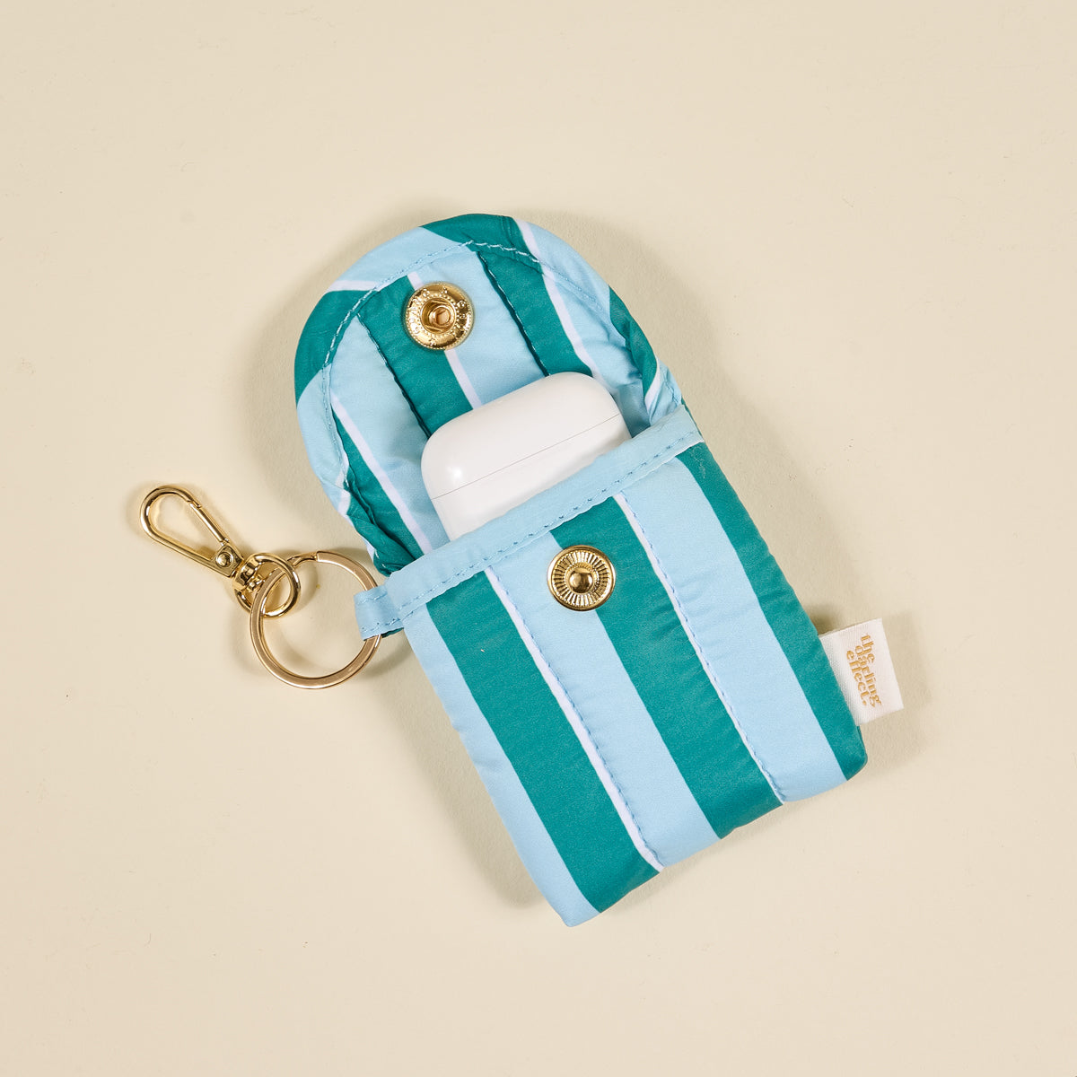The Darling Effect Sicily Stripes Teal Puffy Earbud Case is a blue and turquoise striped, gold-clasped fabric case with a keychain, label, and holds a white wireless earbuds case. Shown against a light beige background.