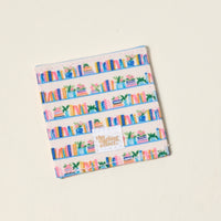The Book Shelfie Corner Keeper Fabric Bookmark by The Darling Effect features a colorful bookshelf pattern with books and plants on beige shelves—a perfect gift for book lovers. A small white tag says "the sharing effect" on an off-white background.