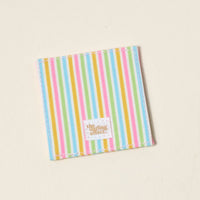 The Color Me Happy Corner Keeper Fabric Bookmark by The Darling Effect features vertical pastel stripes and a white label with gold lettering—an adorable, square corner bookmark and a perfect gift for book lovers.