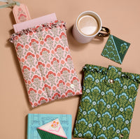 Two floral fabric pouches—one pink, one green—rest on a beige surface beside coffee, two notebooks (blue and green), an envelope with a card, and The Darling Effect's Everbloom Forest Green Corner Keeper Fabric Bookmark—perfect for book lovers.