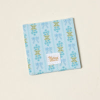 The Oh So Fancy Blue Corner Keeper Fabric Bookmark by The Darling Effect is a square blue coaster with light blue and green floral and bow patterns, plus a small white label—an ideal unique gift for book lovers.