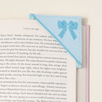 A close-up of an open book with The Darling Effect's Oh So Fancy Blue Corner Keeper Fabric Bookmark, featuring small bows, on a white surface—an ideal gift for book lovers. Some text appears on the left page.