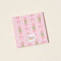 The Oh So Fancy Pink Corner Keeper Fabric Bookmark by The Darling Effect features pink stripes, bows, and flowers on an off-white background with a gold-branded label—an ideal gift for book lovers.