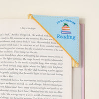 The Darling Effect’s “I’d Rather Be Reading Corner Keeper” fabric bookmark, in blue and yellow with a book stack design, clips to your book’s corner—a perfect gift for book lovers—shown resting on a white surface.