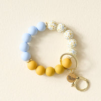 The Darling Effect’s Country Cottage Hands-Free Keychain Wristlet features light blue silicone beads, white floral-patterned beads, and mustard yellow beads, finished with a gold keyring, clasp, and engraved charm.