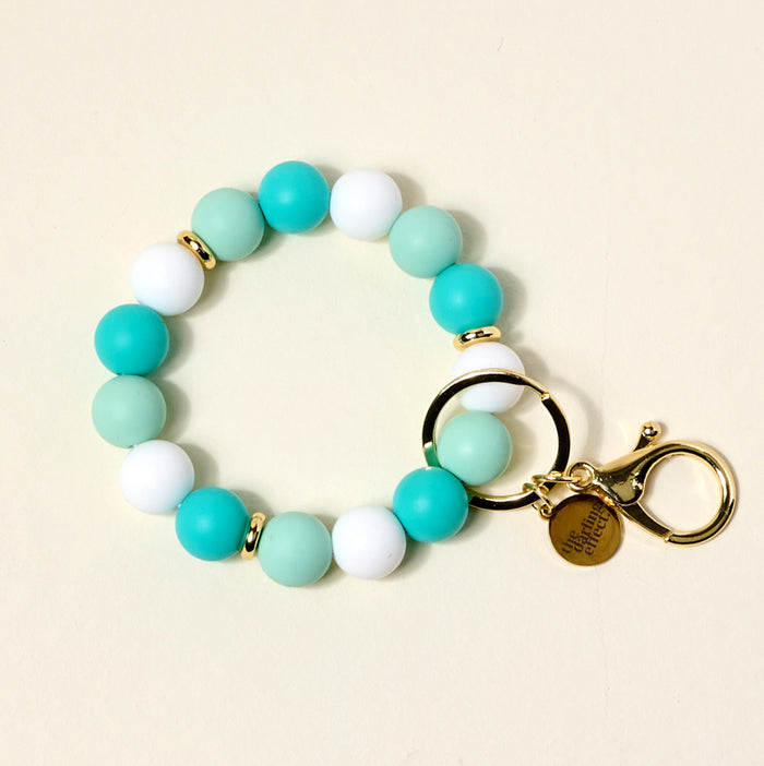 The Darling Effect Daydreamer Hands-Free Keychain Wristlet features alternating turquoise, white, and light green silicone beads, gold spacer beads, a gold keyring, lobster clasp, and a round charm engraved with text.