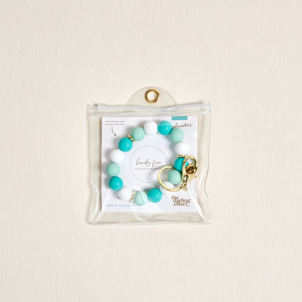 The Darling Effect’s Daydreamer Hands-Free Keychain Wristlet features teal and white silicone beads with a gold keyring, packaged in a clear pouch against a light beige background—stylish convenience for on-the-go use.