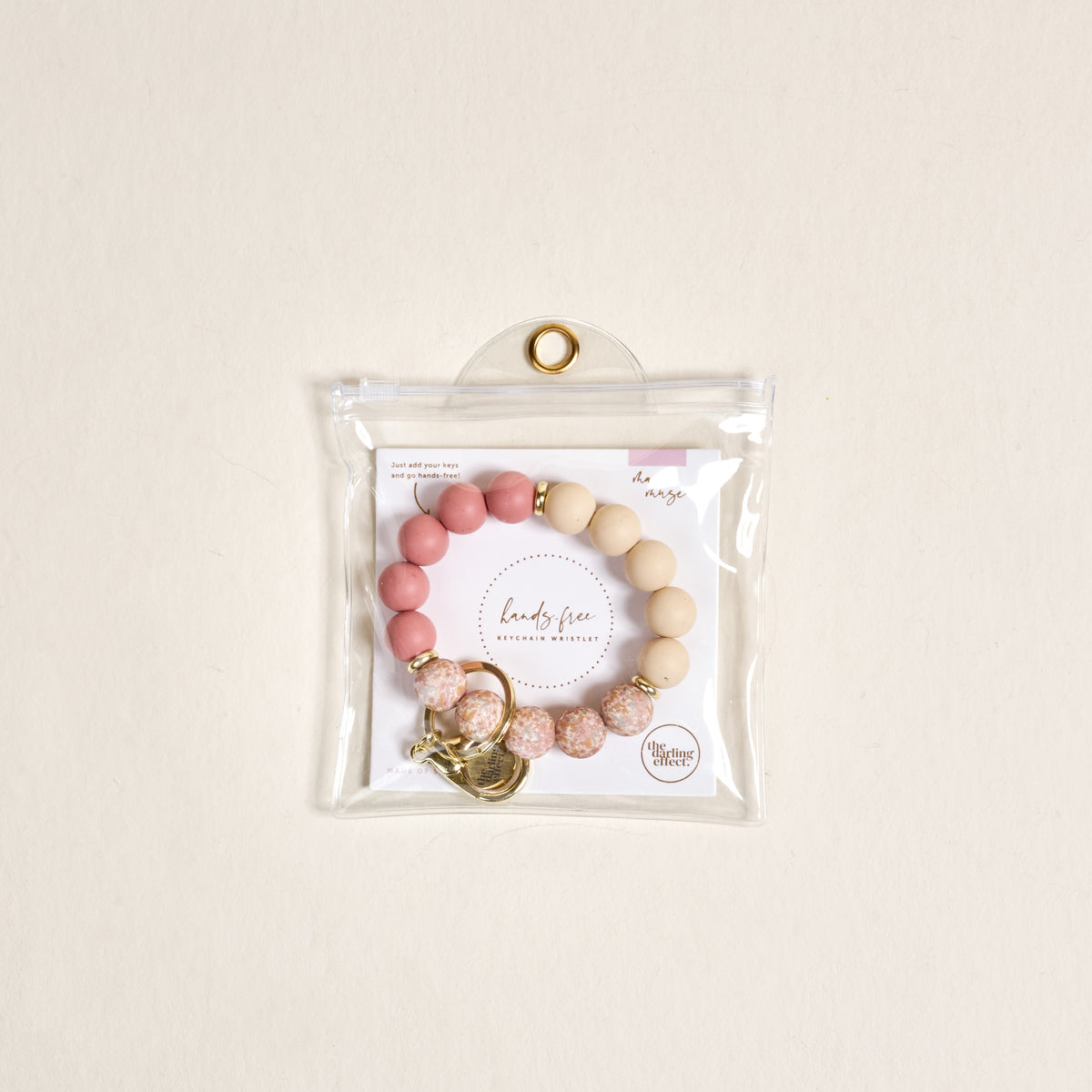 The-darling-effect’s Mauve Muse Hands-Free Keychain Wristlet, featuring pink and beige silicone beads with gold accents, is shown in a clear pouch with a gold grommet against a light beige background.