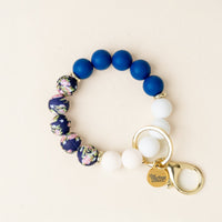 The-darling-effect Notes of Navy Hands-Free Keychain Wristlet features blue, white, and floral silicone beads, a gold clasp, and a gold tag with the brand’s name, shown on a cream background for easy hands-free use.
