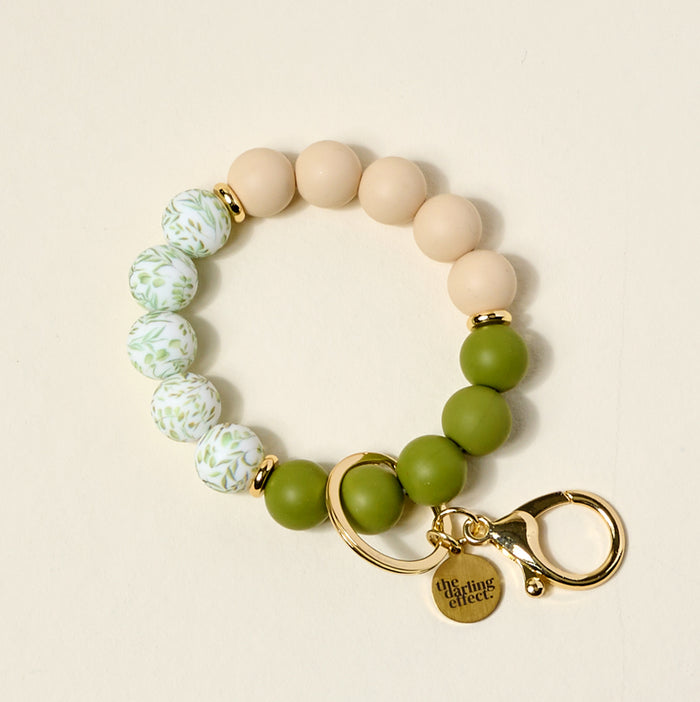 The Olive Branch Hands-Free Keychain Wristlet by The Darling Effect features beige, green, and white silicone beads with gold accents, a keyring, lobster clasp, and a gold charm engraved with the brand name for stylish hands-free carrying.