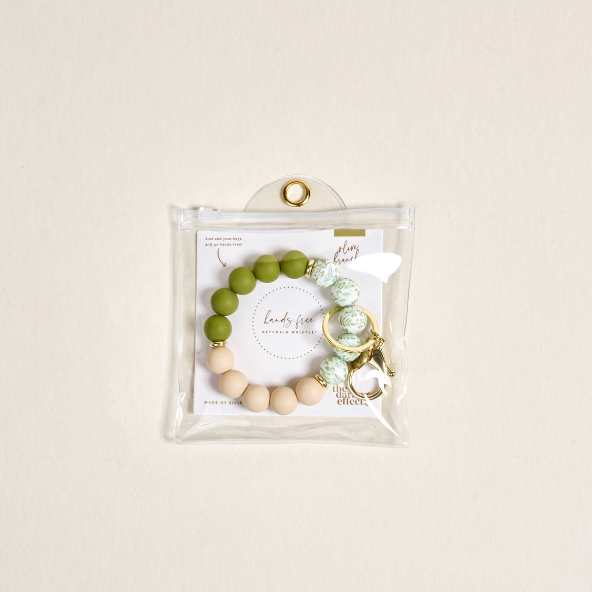 The Darling Effect’s Olive Branch Hands-Free Keychain Wristlet features green, white, and beige silicone beads with a gold keyring, packaged in a clear snap pouch and includes a branded card for extra style.