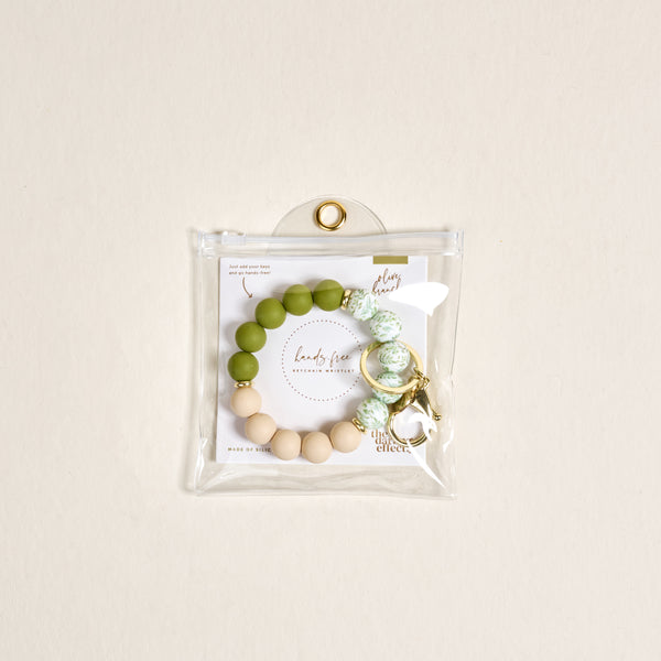 The Darling Effect’s Olive Branch Hands-Free Keychain Wristlet features green, white, and beige silicone beads with a gold keyring, packaged in a clear snap pouch and includes a branded card for extra style.