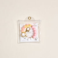 The Darling Effect’s Pink Lemonade Hands-Free Keychain Wristlet features pink, yellow, and white silicone beads with a gold keyring and clasp, shown in a clear pouch on a light beige background for stylish convenience.