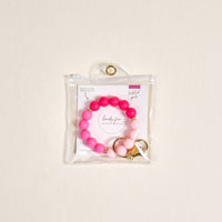 The Darling Effect Tickled Pink Hands-Free Keychain Wristlet, a beaded pink and white silicone key holder with gold accents, is shown in a clear pouch with a gold grommet. The pouch includes an insert reading "lady flow" and "Twisted pink.