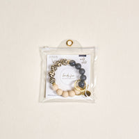 The Wild Child Hands-Free Keychain Wristlet by the-darling-effect features leopard print, dark gray, and beige silicone beads with gold key rings and charms, all in a clear button-closure pouch for chic, hands-free carrying.