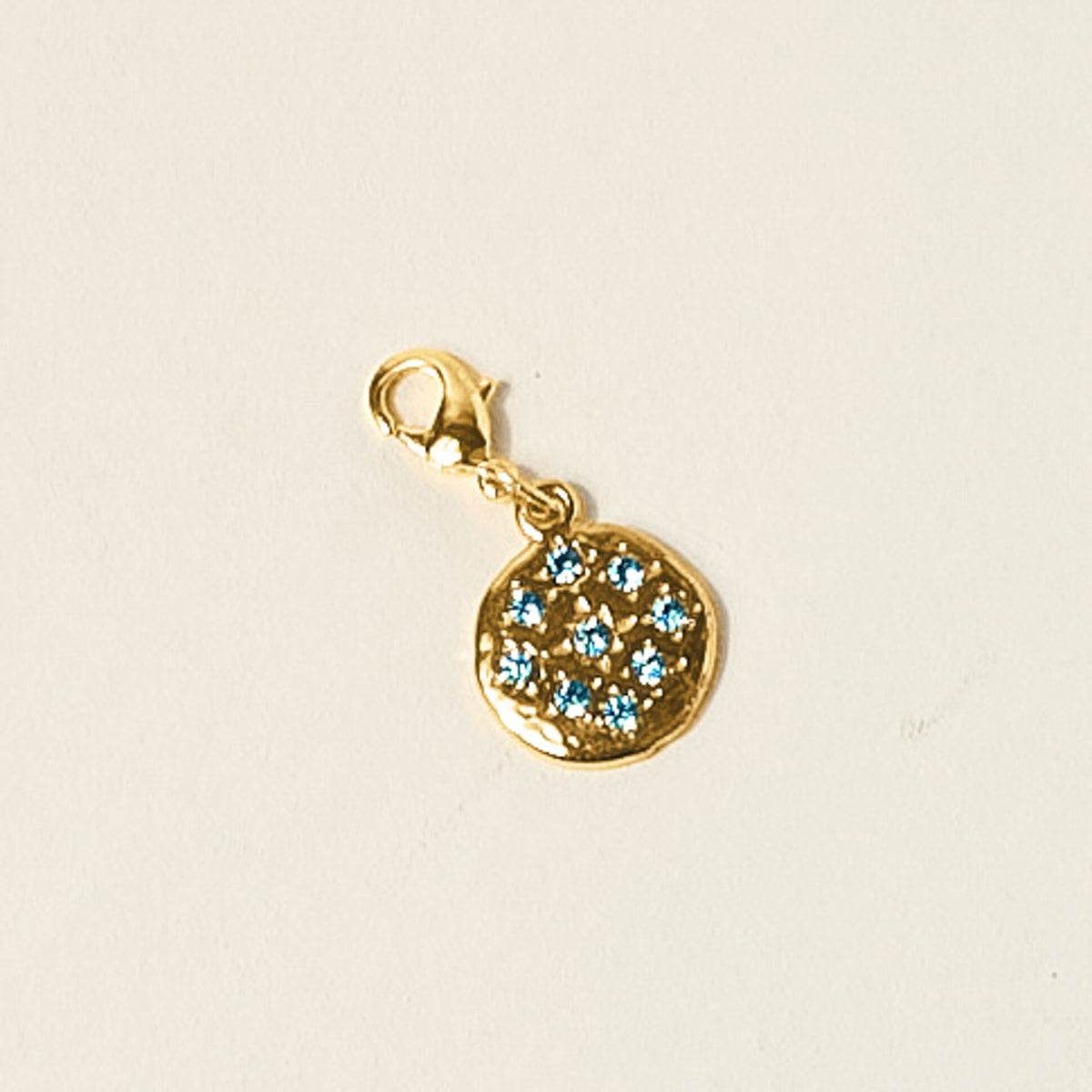 The Darling Effect Blue Jewels Jewelry Charm Gold features a round, textured gold pendant with small blue gemstones in a circular design and a lobster clasp—ideal for customizing your charm jewelry or favorite necklace.