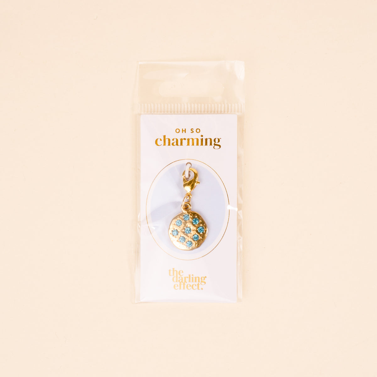 The Darling Effect Blue Jewels Jewelry Charm Gold features blue and white gems in a circular design, attached to a clasp and packaged on an OH SO charming card inside a clear bag—perfect for your charm necklace or unique collection.