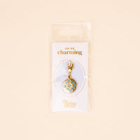 The Darling Effect Blue Jewels Jewelry Charm Gold features blue and white gems in a circular design, attached to a clasp and packaged on an OH SO charming card inside a clear bag—perfect for your charm necklace or unique collection.