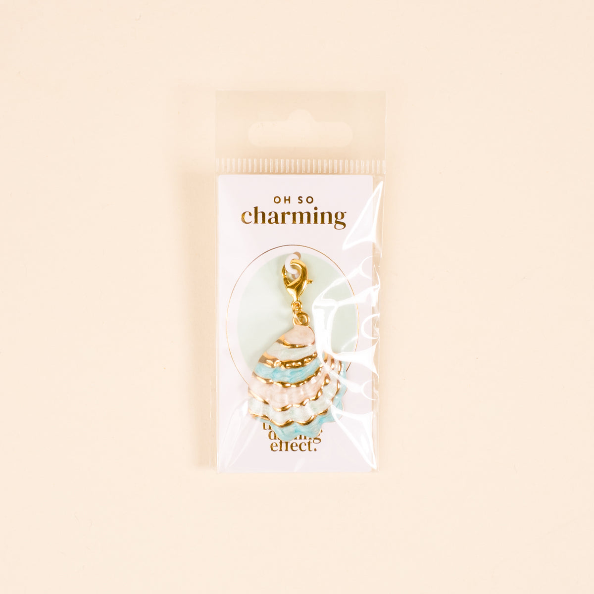 The Darling Effect’s Blue Shell Beauty Jewelry Charm Gold, shaped like a seashell with turquoise enamel, is shown in clear packaging labeled “Oh So Charming” against a pale background.
