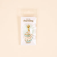The Darling Effect’s Blue Shell Beauty Jewelry Charm Gold, shaped like a seashell with turquoise enamel, is shown in clear packaging labeled “Oh So Charming” against a pale background.