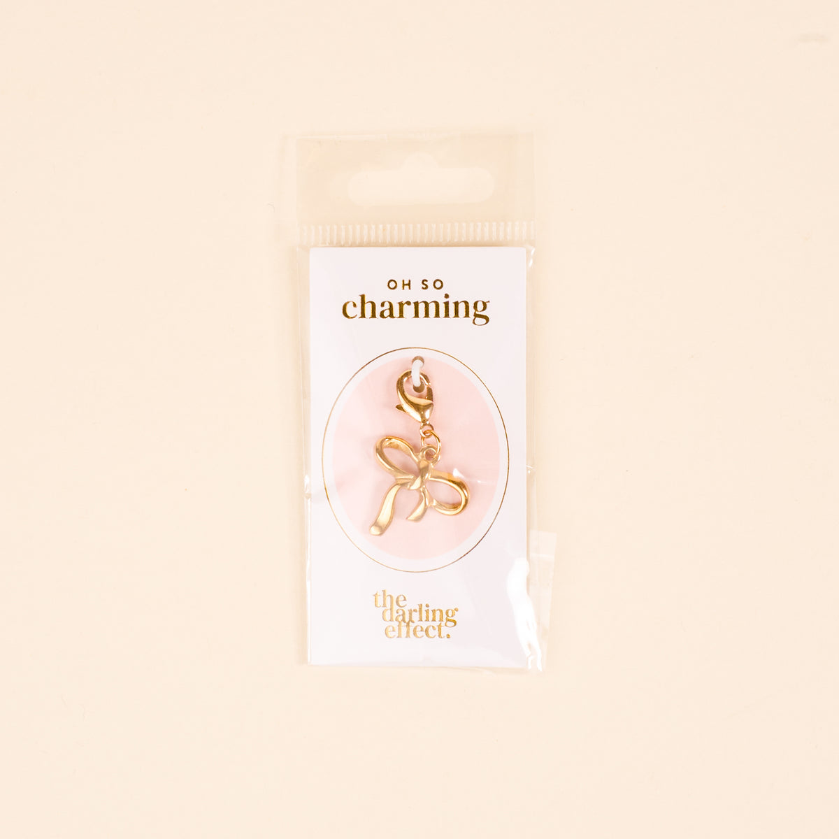 The Bow Jewelry Charm Gold by the-darling-effect is shown in clear packaging on a pink background. The text reads "OH SO charming" and "the darling effect," making it perfect for charm jewelry or cute necklaces.