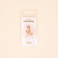 The Bow Jewelry Charm Gold by the-darling-effect is shown in clear packaging on a pink background. The text reads "OH SO charming" and "the darling effect," making it perfect for charm jewelry or cute necklaces.
