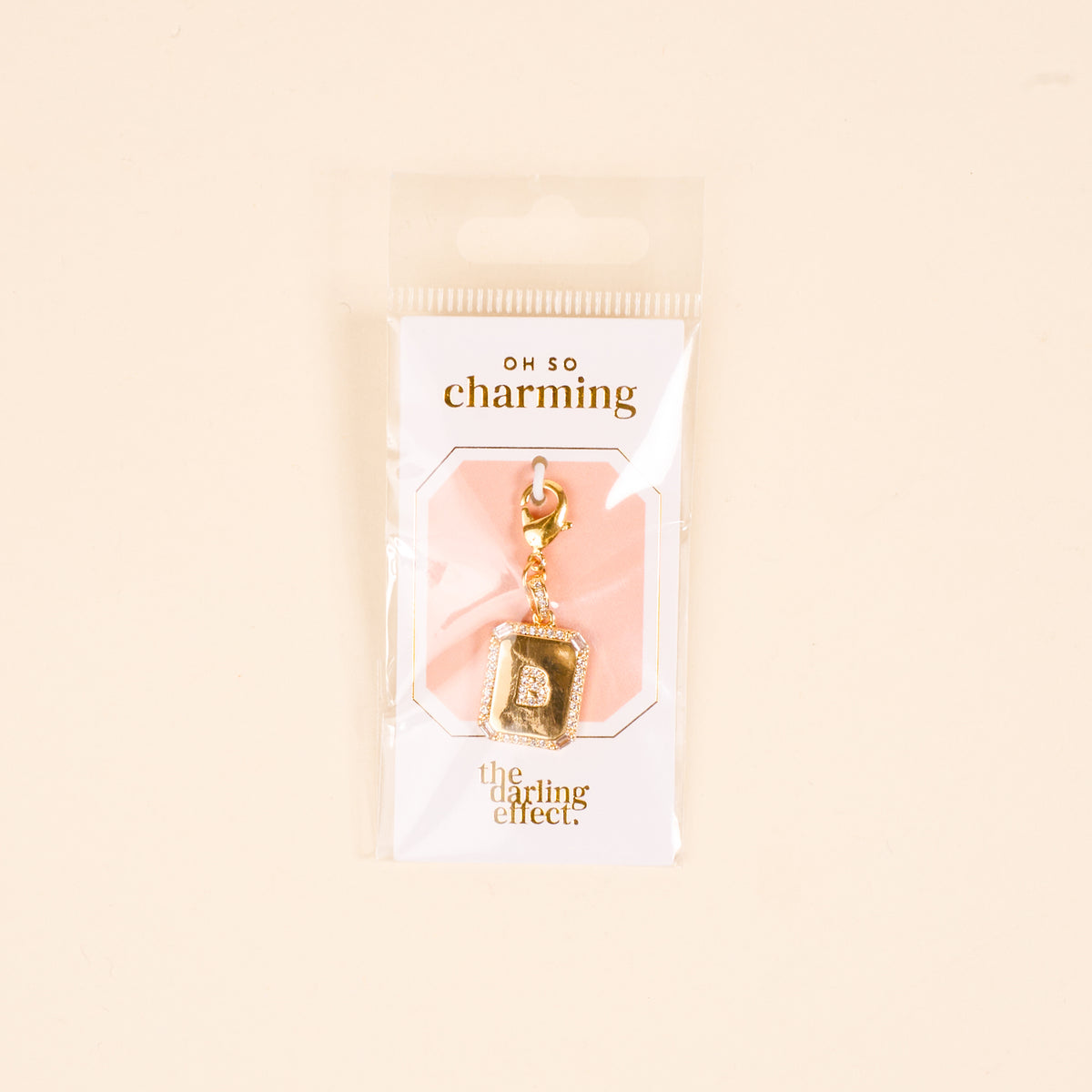 The-darling-effect B Jewelry Charm Initial Letter Gold features a gold-plated "B" charm on a white and peach card with "oh so charming" and "the darling effect," packaged in a clear plastic bag.