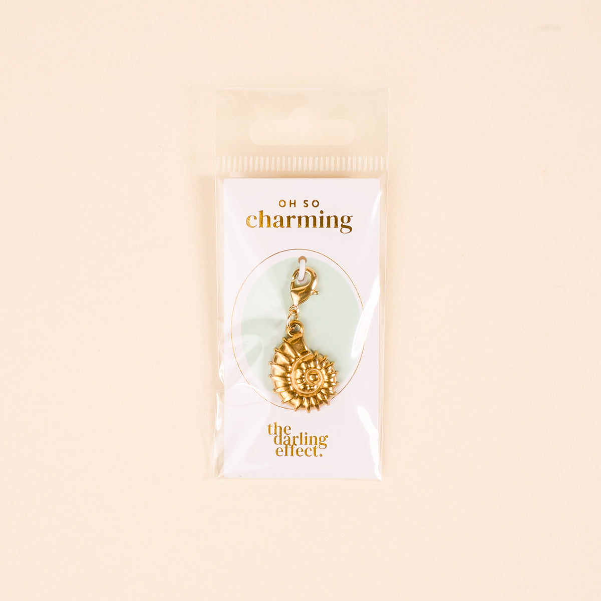 The Darling Effect's Conch Shell Jewelry Charm Gold features a clasp and comes on a white card reading "OH SO charming" and The Darling Effect, packaged in a clear bag with a light beige background—ideal for your charm bar or unique jewelry.