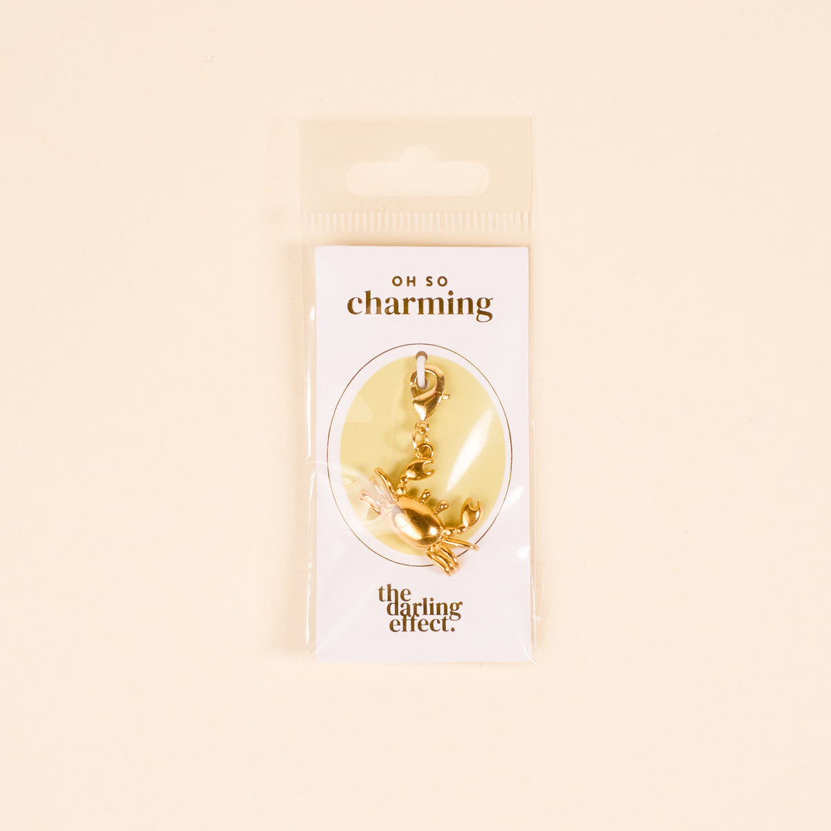 The Darling Effect’s Crab Jewelry Charm Gold hangs from a clasp on a white “OH SO charming” card, perfect for your charm bar or jewelry collection. The card comes in clear plastic packaging.