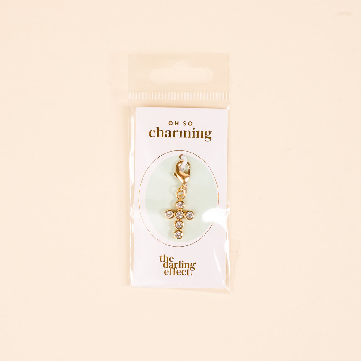 The Cross Jewelry Charm Gold by the-darling-effect features sparkling rhinestones and is attached to an OH SO charming card, packaged in clear plastic against a light beige background—perfect for charm jewelry.