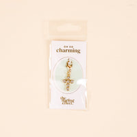 The Cross Jewelry Charm Gold by the-darling-effect features sparkling rhinestones and is attached to an OH SO charming card, packaged in clear plastic against a light beige background—perfect for charm jewelry.