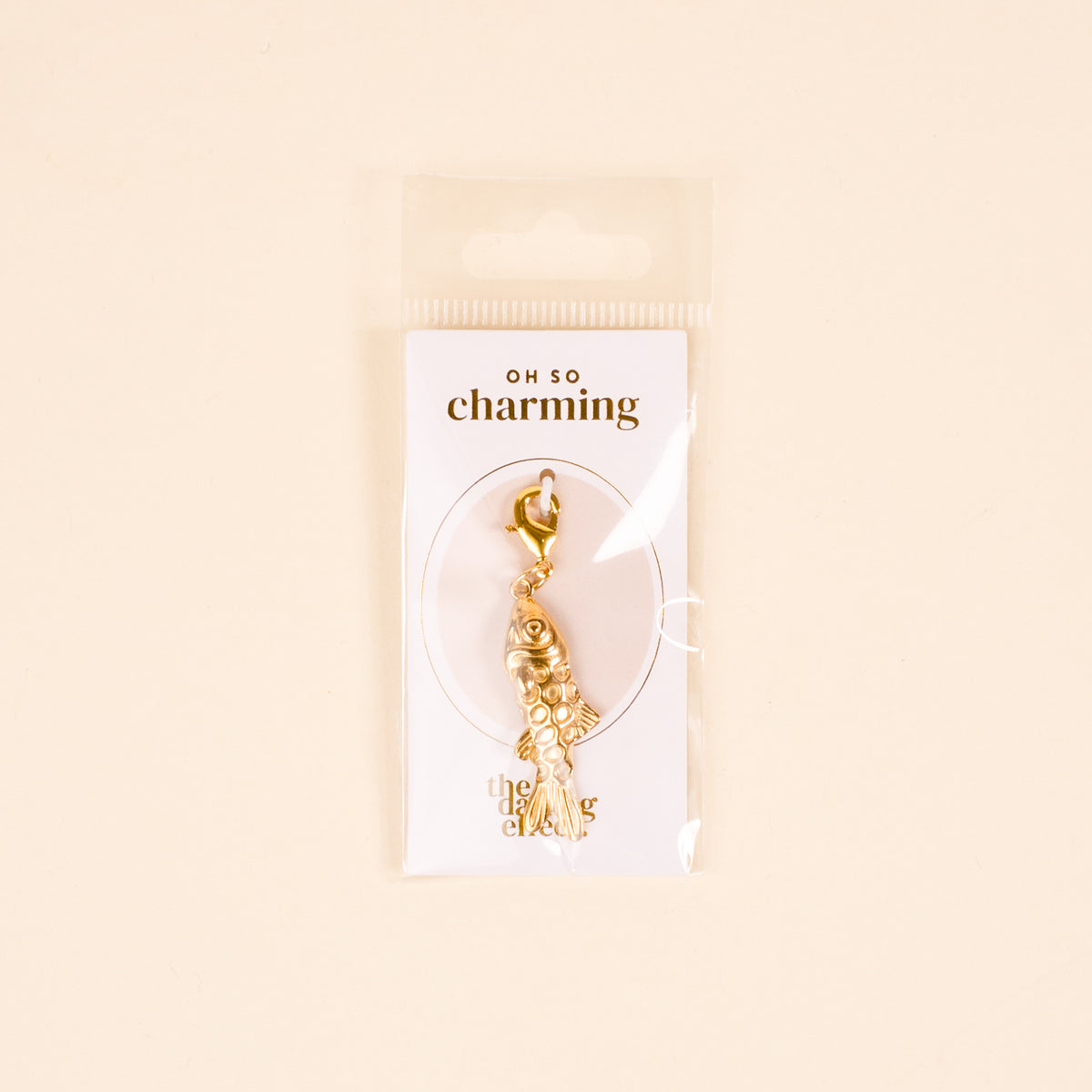 The Darling Effect Fish Jewelry Charm Gold features intricate detailing, comes attached to an "Oh So Charming" card, and is packaged in a clear sleeve against a light beige background—perfect for your charm bar.