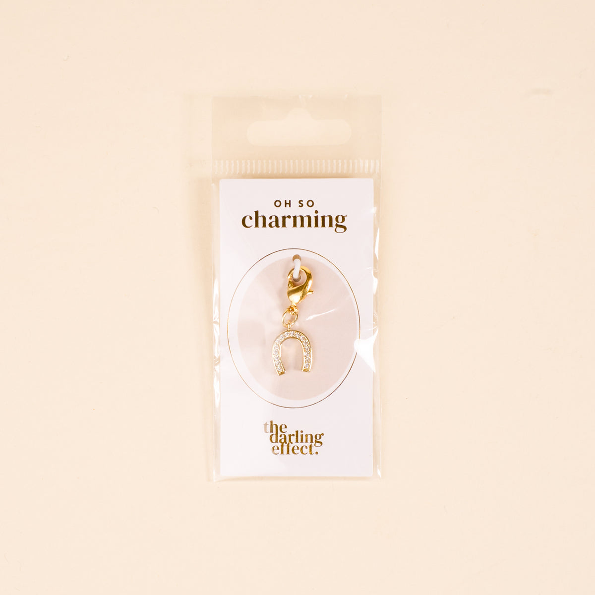 The-darling-effect Horseshoe Jewelry Charm Gold features small crystals, attached to an "OH SO charming" card—perfect for your charm bar. Unique jewelry packaged in a clear plastic bag against a beige background.