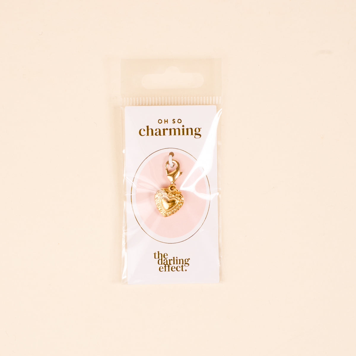 The Heart Jewelry Charm Gold by the-darling-effect features a gold heart-shaped charm with a clasp, displayed on a white Oh So Charming card and presented in clear packaging against a beige background.