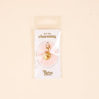 The Heart Jewelry Charm Gold by the-darling-effect features a gold heart-shaped charm with a clasp, displayed on a white Oh So Charming card and presented in clear packaging against a beige background.