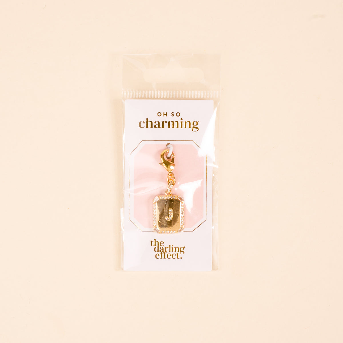 The-darling-effect J Jewelry Charm Initial Letter Gold features a gold-plated brass "J" with a glitter border, clasp, and is displayed on a branded card in clear packaging—perfect personalized jewelry on a cute pink-and-white background.