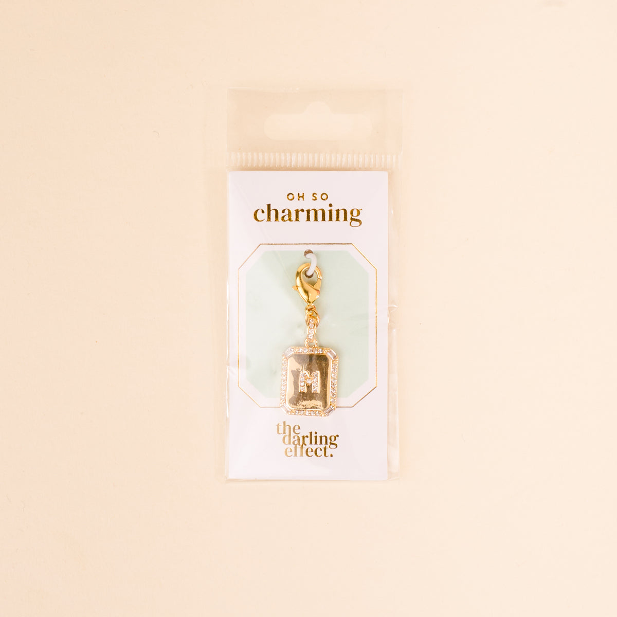 The M Jewelry Charm Initial Letter Gold by the-darling-effect features a gold-plated charm attached to a lobster clasp, displayed on white Oh So Charming packaging—an ideal gift for personalized jewelry lovers.