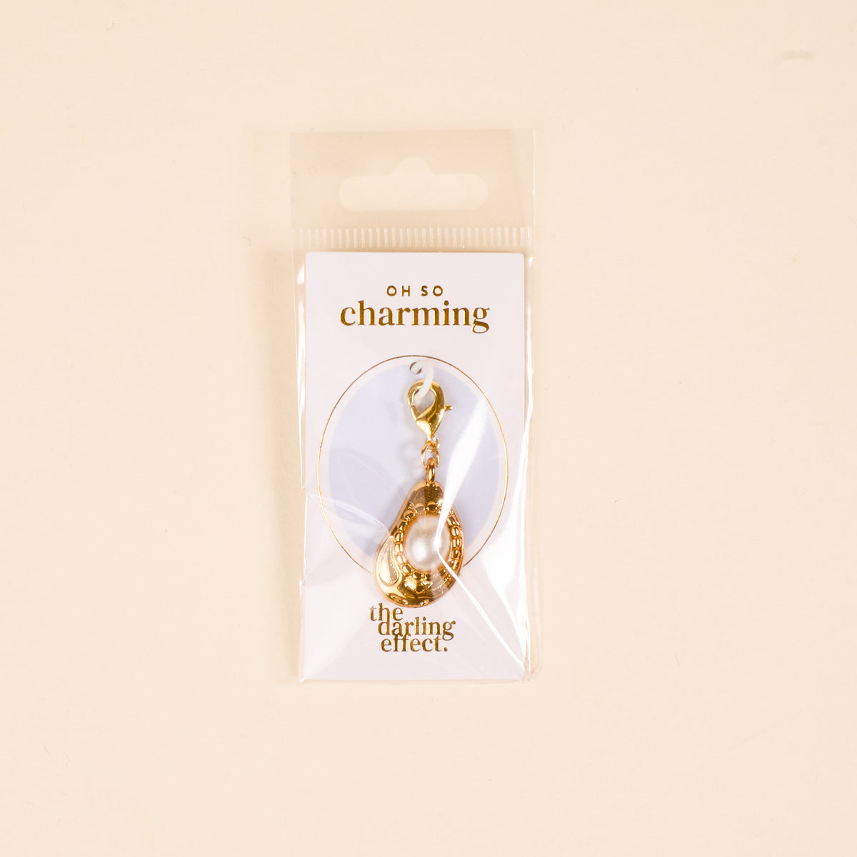 The Darling Effect's Oyster Jewelry Charm Gold keychain features a pearl charm and comes attached to an “OH SO charming & the darling effect” card, all packaged in a clear plastic bag.