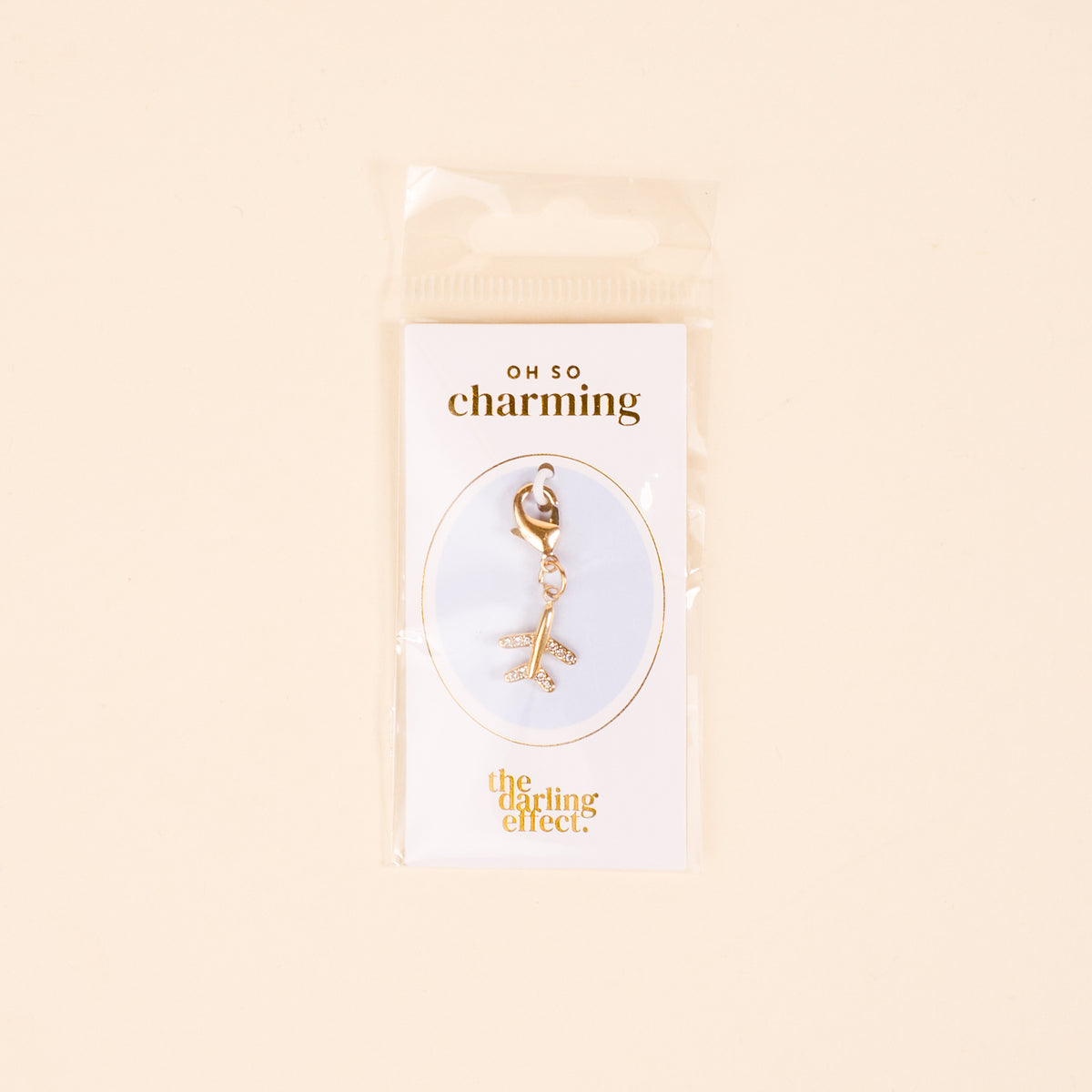 The Plane Jewelry Charm Gold by the-darling-effect features a gold airplane on a lobster clasp, displayed on a white card with gold "oh so charming" and "the darling effect" text, and comes packaged in a clear plastic bag.