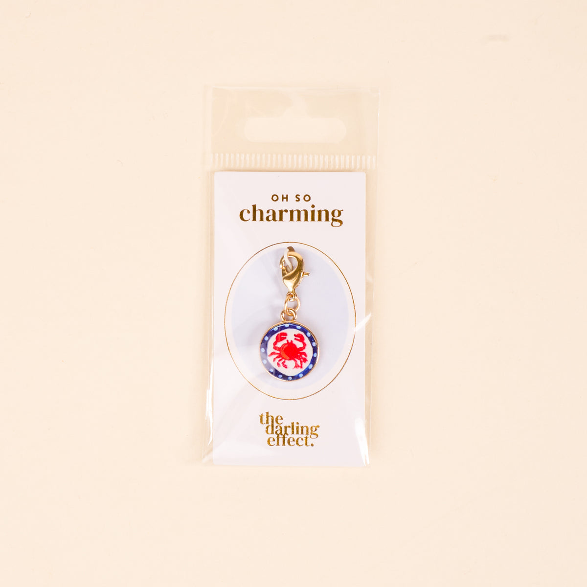 The Darling Effect Painted Crab Jewelry Charm Gold features a red crab on a blue and white background with a gold clasp, perfect for your charm bar. Packaged on a white Oh So Charming card, it’s a delightful jewelry addition.