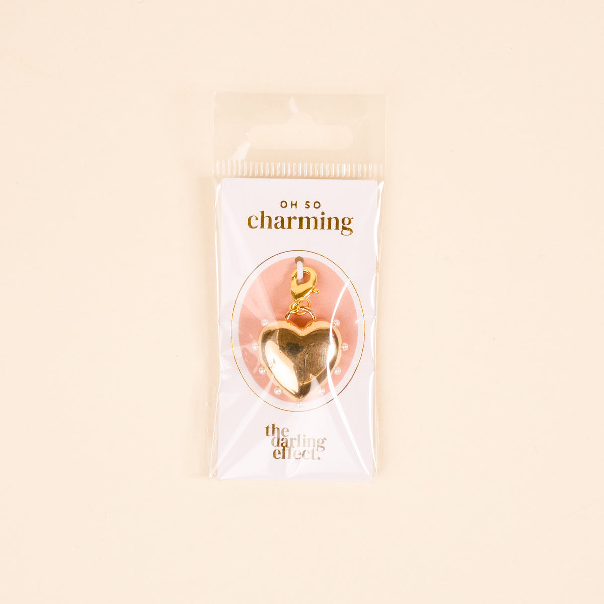 The Darling Effect’s Pearl Lined Heart Jewelry Charm Gold features a lobster clasp, ideal for layering on your charm bar. It comes on a white and pink OH SO charming card in a clear bag against a pale beige background.